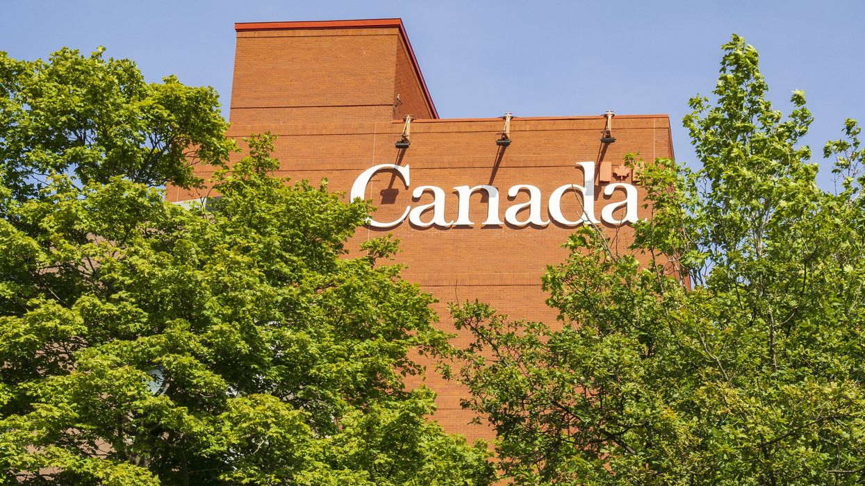 Red brick building with lettering reading "Canada," with trees in the foreground and a clear blue sky behind.