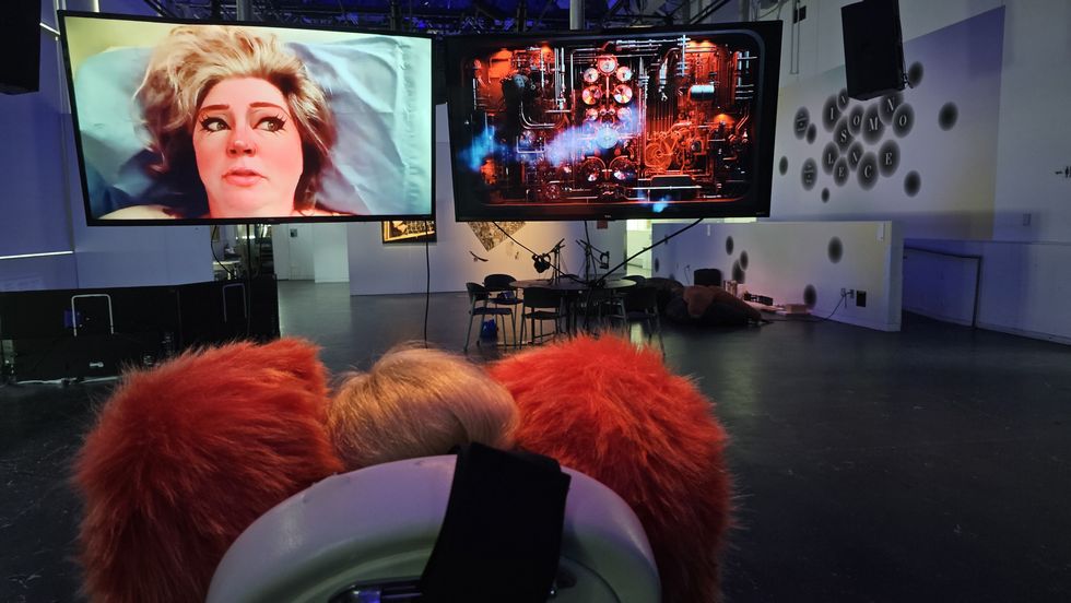 Sofia faces two screens wearing a pair of large fluffy red headphones and watches an AI version of Montreal artist Dayna McLeod, next to a shot of a machine emitting smoke.