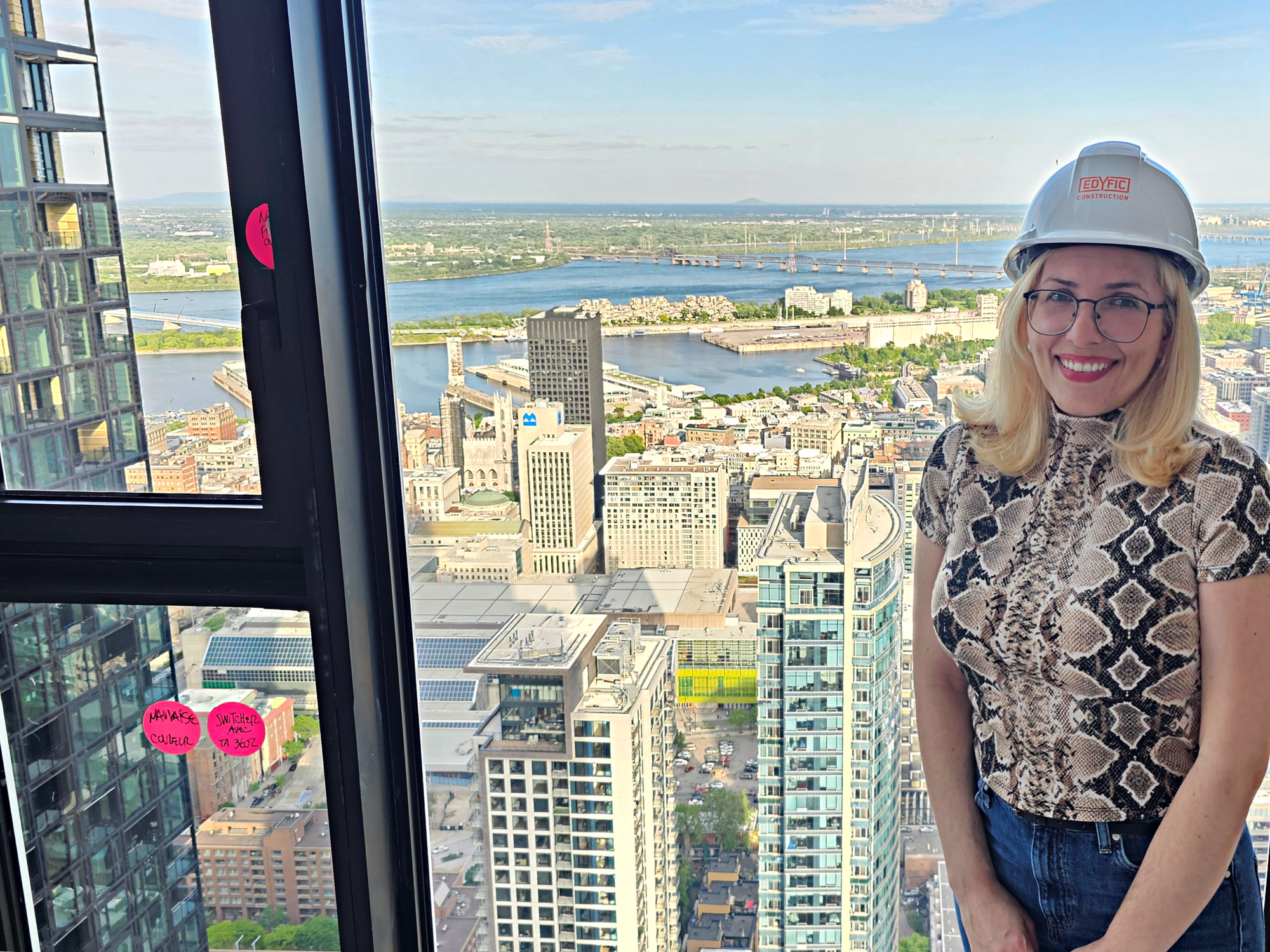 Sofia wears a hardhat in front of a penthouse window overlooking the south side of downtown Montreal.