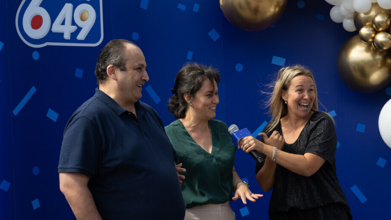 Someone holds a microphone up to two people in front of a blue Lotto 6/49 backdrop.