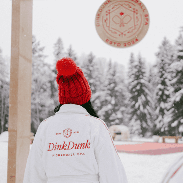 Someone in a branded DinkDunk bathrobe looks at a pickleball court surrounded by snow.