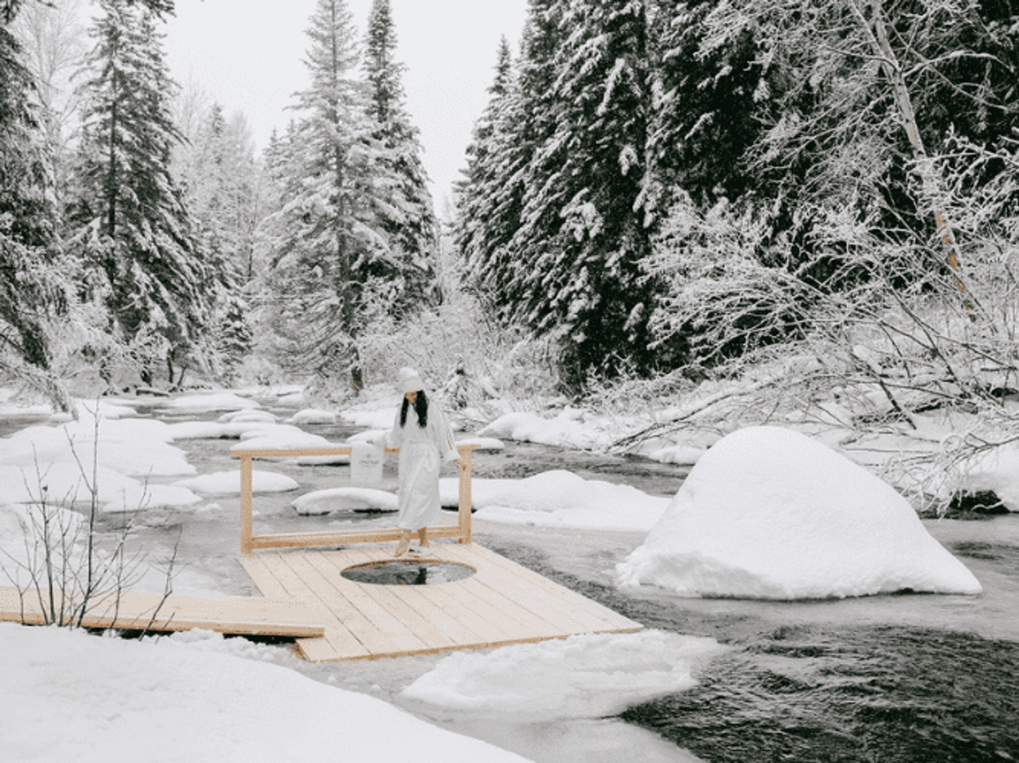Someone in a white bathrobe and hat dips a toe in a cold plunge bath on a platform floating in a snowy river.