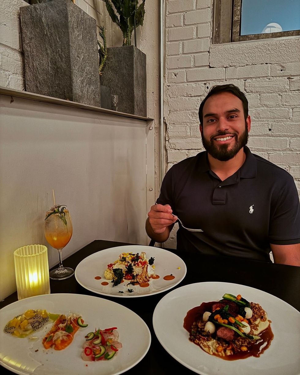 Someone sits smiling at a restaurant table with a fork in hand, a cocktail and plates of food in front of them.