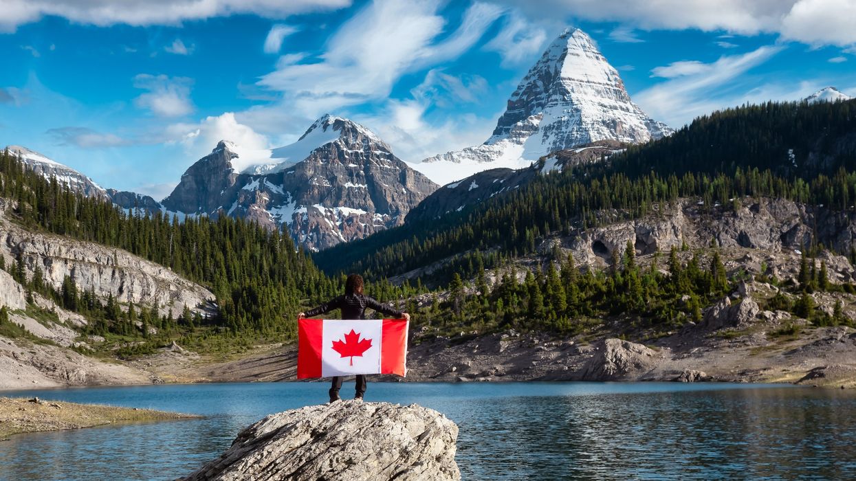 Someone standing in front of a lake holding a Canadian flag with the Canadian Rockies in the background (illustrative).