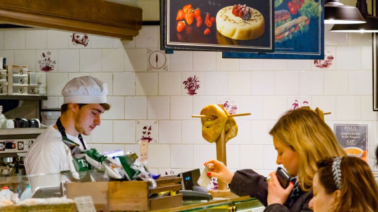 Someone taps their credit card on a pay terminal at a café., while a person wearing a chef hat stands behind the counter.