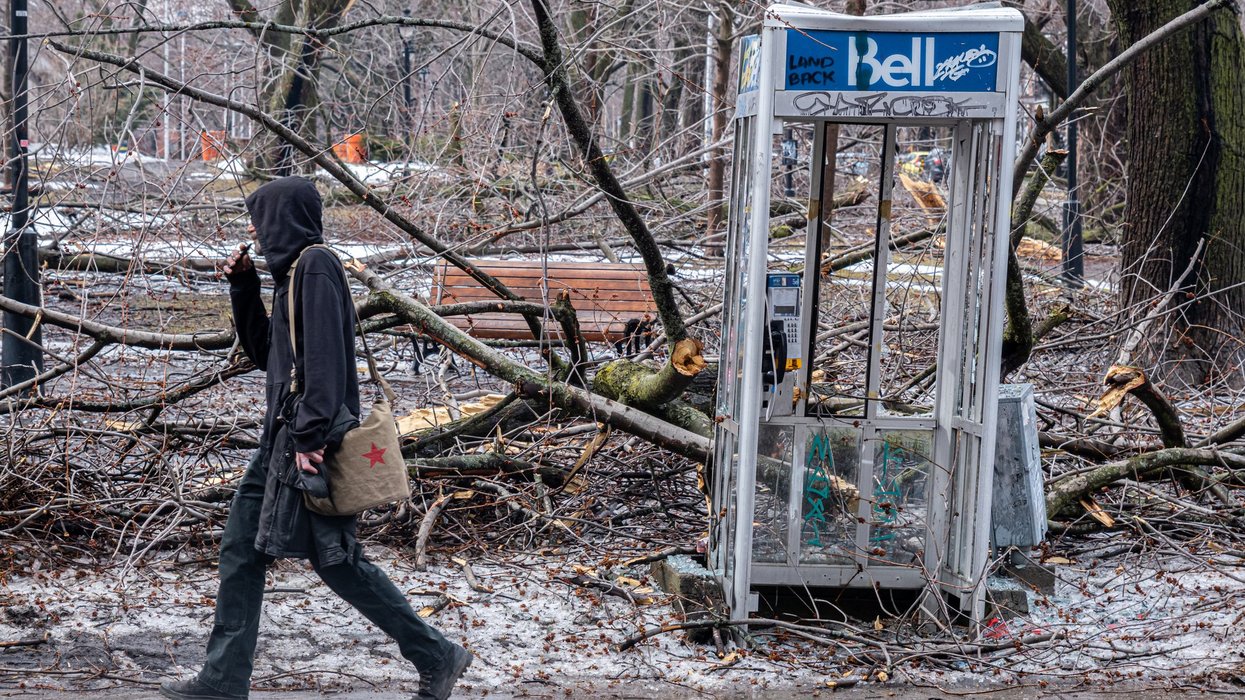 Someone walks by a damaged telephone booth with icy fallen trees scattered around it.