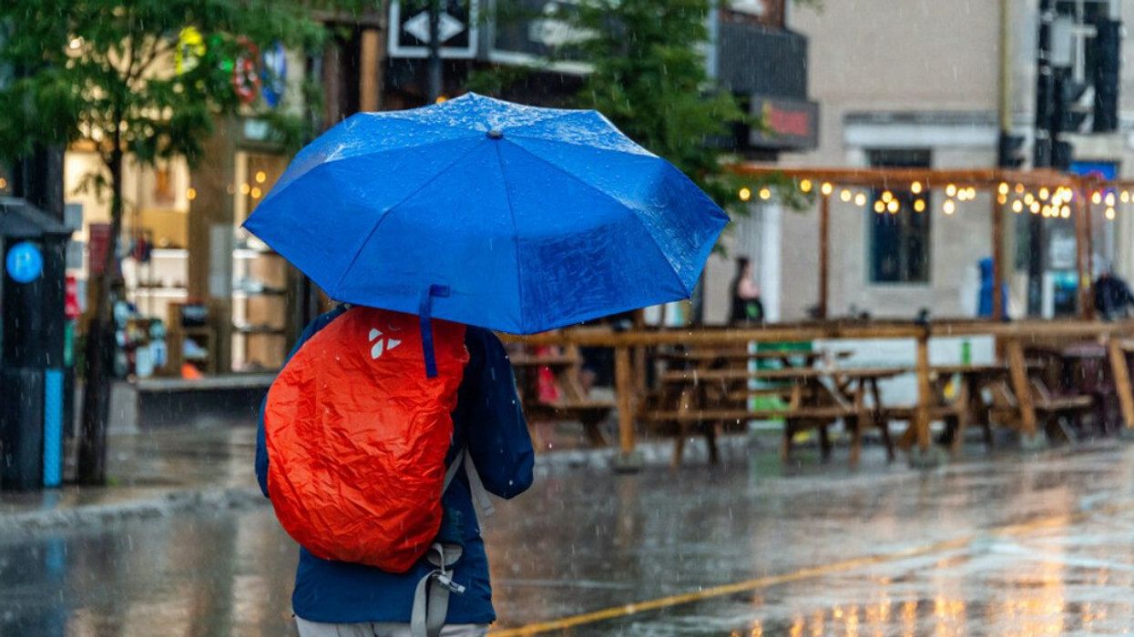 Someone wearing a red backpack holds up a bright blue umbrella as they pass by a Mont Royal terrasse on a rainy day.
