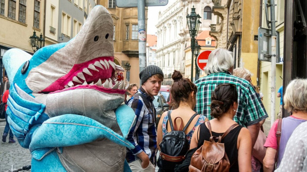 Someone wears a giant shark costume in the street as people pass.