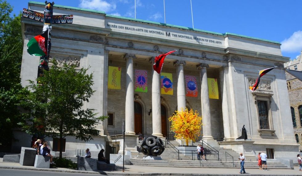 The exterior of Montreal's museum of fine arts with three flags flying on the rooftop and various art installations located in the front of the museum.