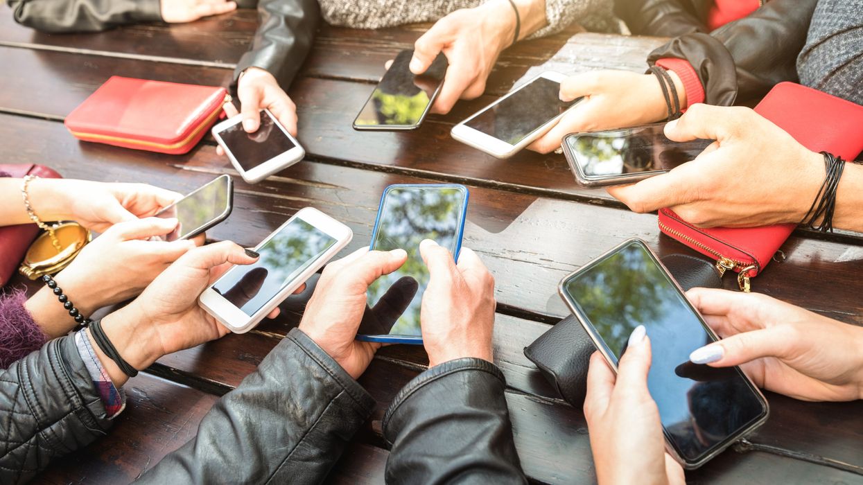 The hands of eight people holding different models of smartphone over a brown wooden outdoor table.