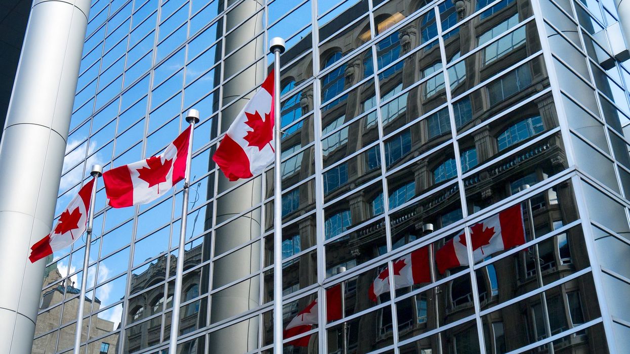 Three Canada flags blowing in the wind with their reflections against a mirrored window office building.