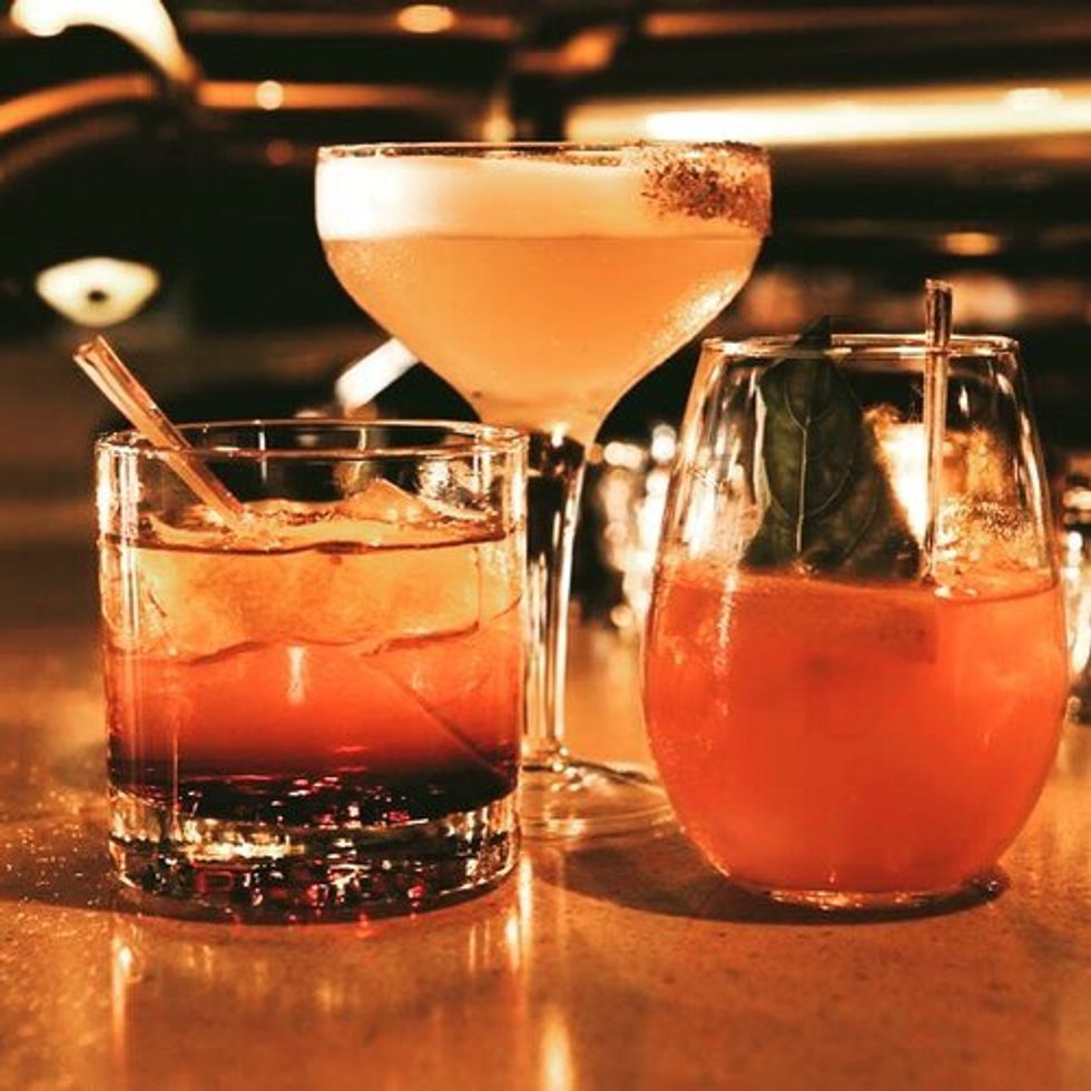 Three colorful cocktails on a bar counter, featuring a dark whiskey drink with an ice cube, a light yellow cocktail in a coupe glass with a sugared rim, and a vibrant orange drink garnished with a basil leaf in a stemless glass.