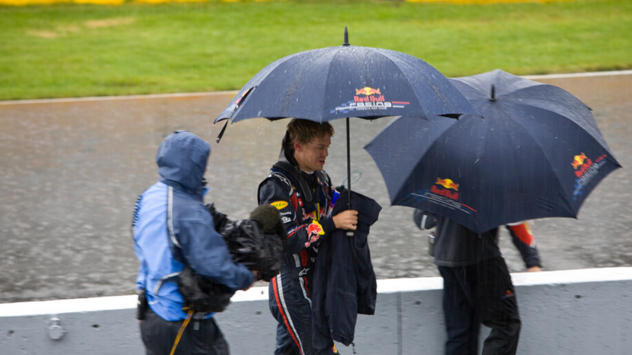 Three people stand on Montreal's Circuit Gilles-Villeneuve, two of whom wear race car driving suits and hold umbrellas.