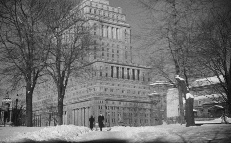 Two figures walk down a snowy path in Dorchester Square in front of the Sun Life Building.