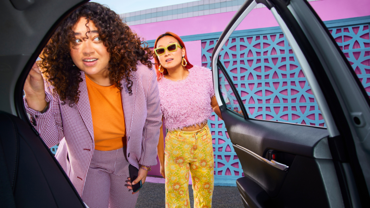 Two people in colourful outfits peer into the passenger seat of a car through an open door.