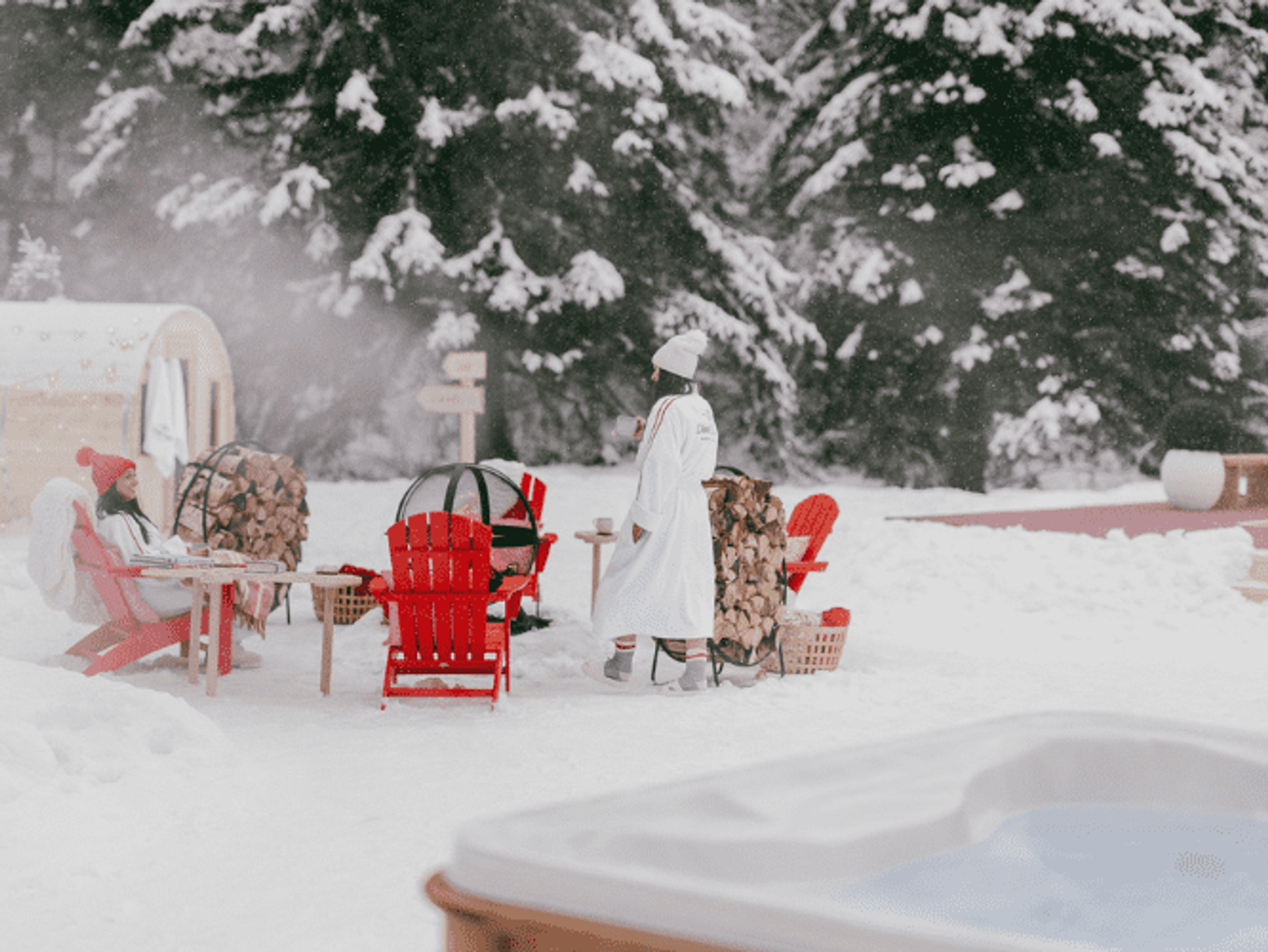 Two people in white bathrobes, tuques and socks near a fire pit in a snowscape featuring a barrel sauna, outdoor jacuzzi and pickleball court.