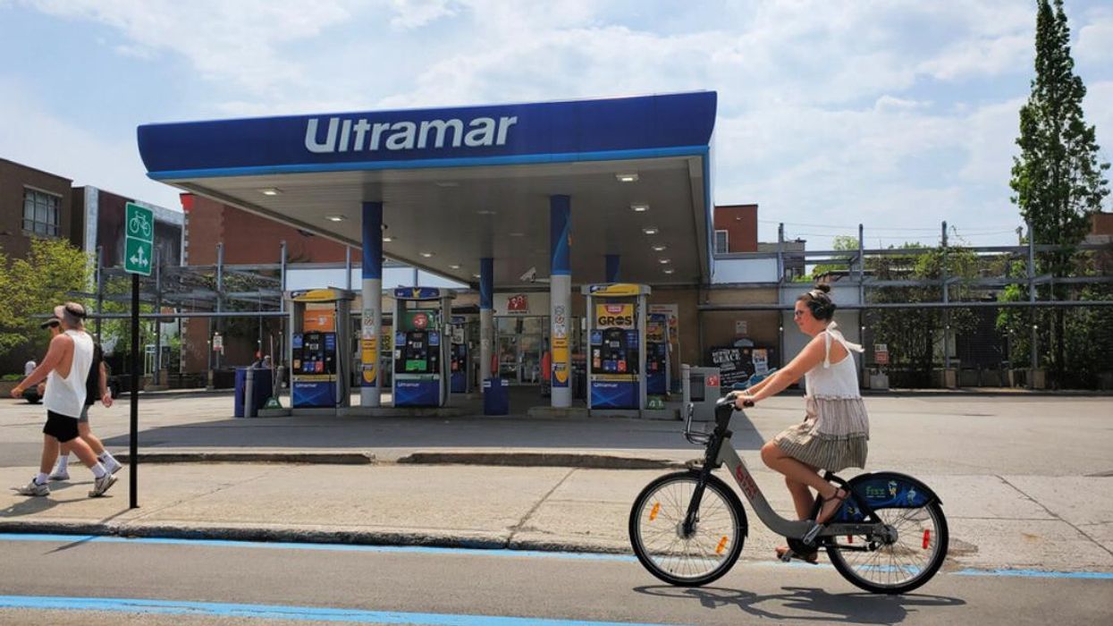 Two people walk by an Ultramar gas station, while another person bikes past.