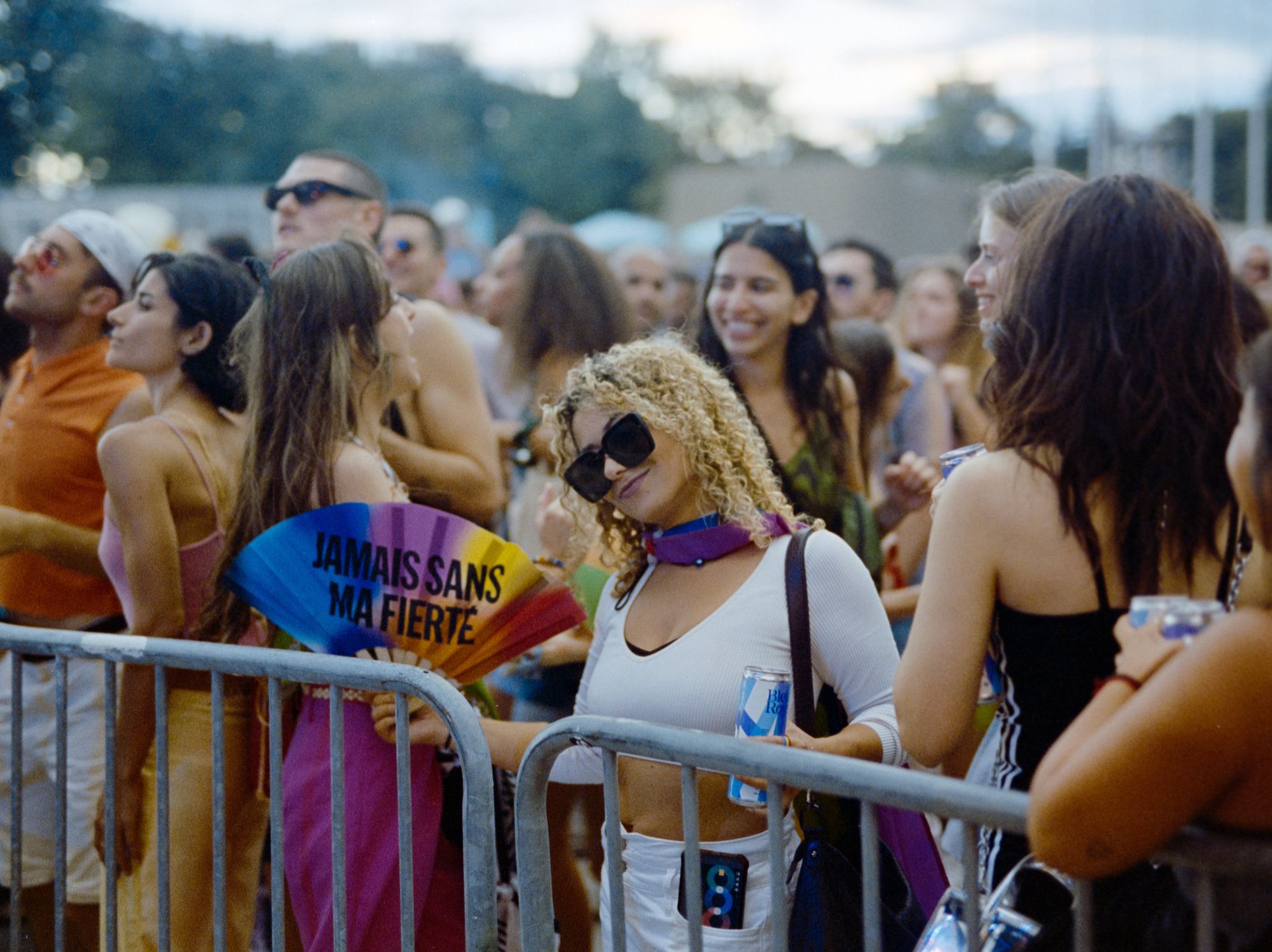 \u200bA person at Montreal Pride wearing sunglasses and a rainbow flag scarf holds up a multi-coloured fan that reads "Jamais sans ma fiert\u00e9" (Never without my pride.)