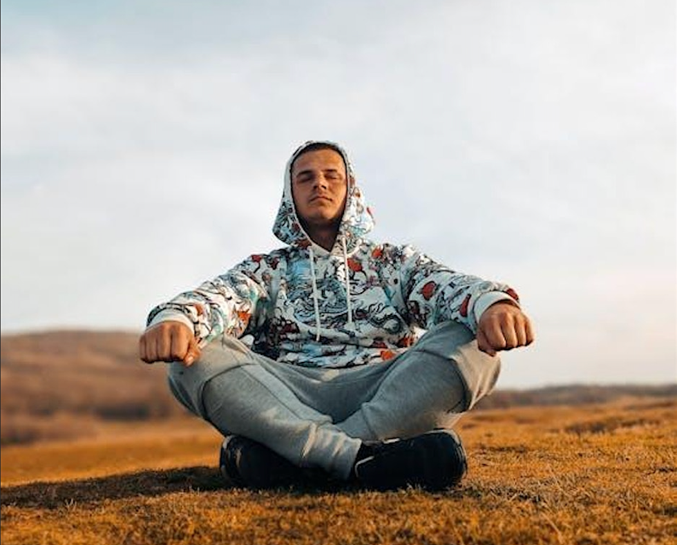 \u200bA person sitting on the ground with their leg crossed and eyes closed in a meditative stance.