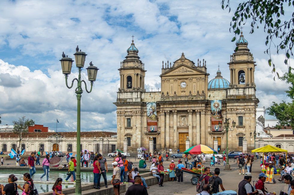 \u200bPeople sit around a fountain in front of a cathedral in Plaza de la Constitucion Constitution Square in Guatemala City.