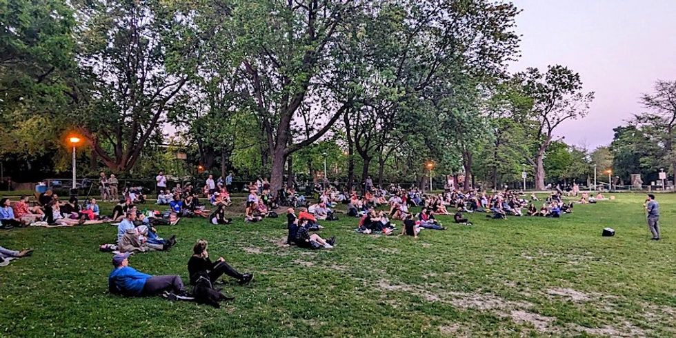 \u200bPeople sitting in a Montreal park watching a stand-up comedy show performance.