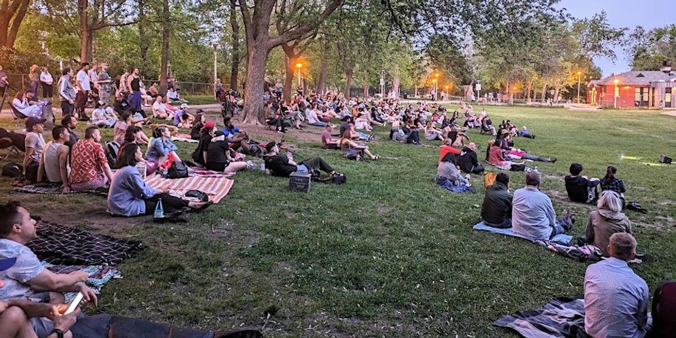 \u200bPeople sitting in a park watching someone perform stand-up comedy.