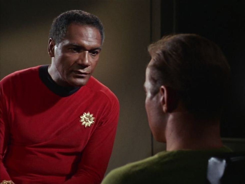 \u200bPercy Rodriguez in a red costume with a gold sun emblem, as Commodore Stone in "Star Trek."