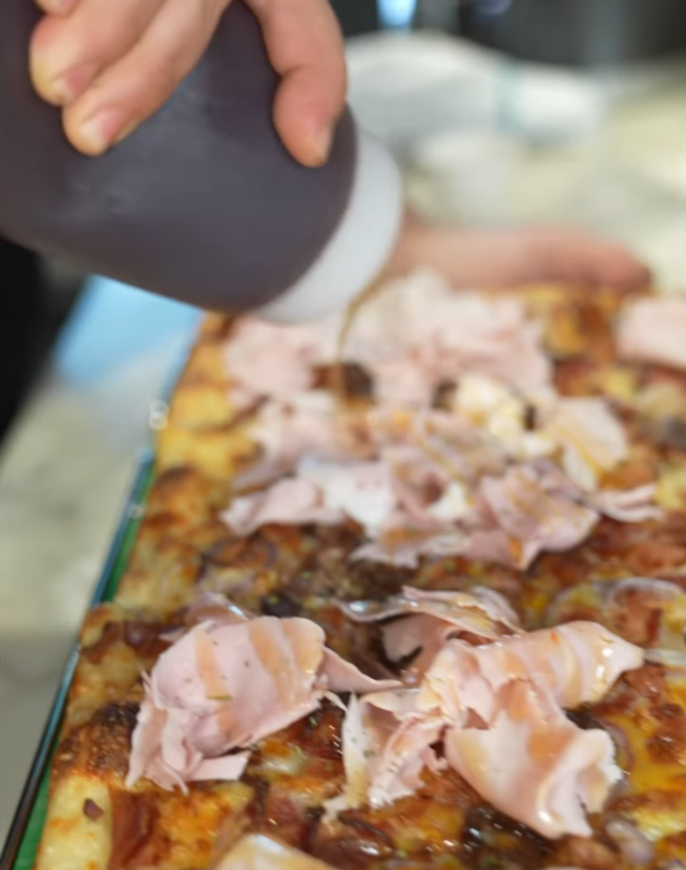 \u200bSomeone drizzles maple syrup on pizza topped with prosciutto, bacon, and onions.