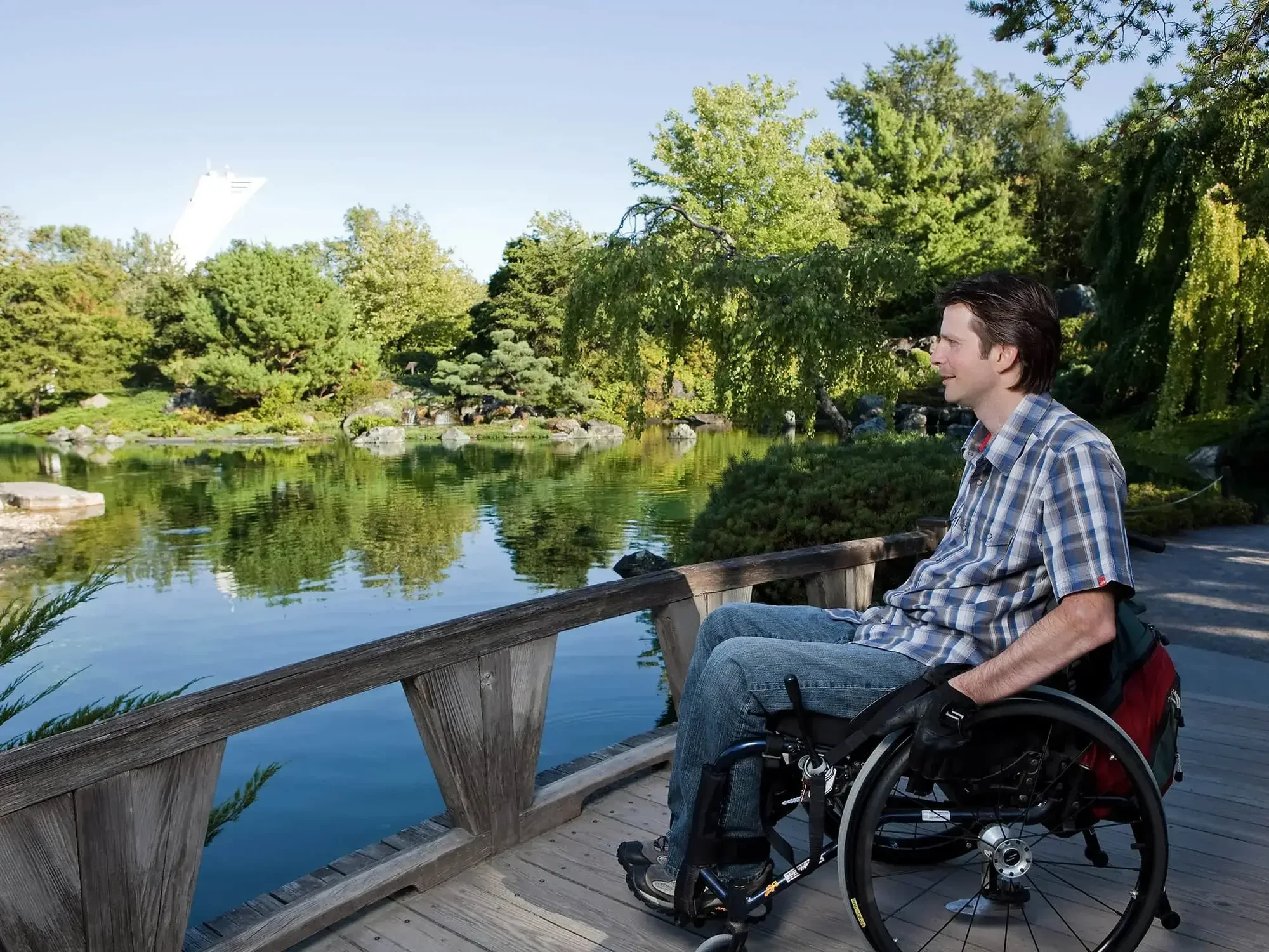 \u200bSomeone in a wheelchair on a wooden bridge overlooked a lake and the Olympic Tower.