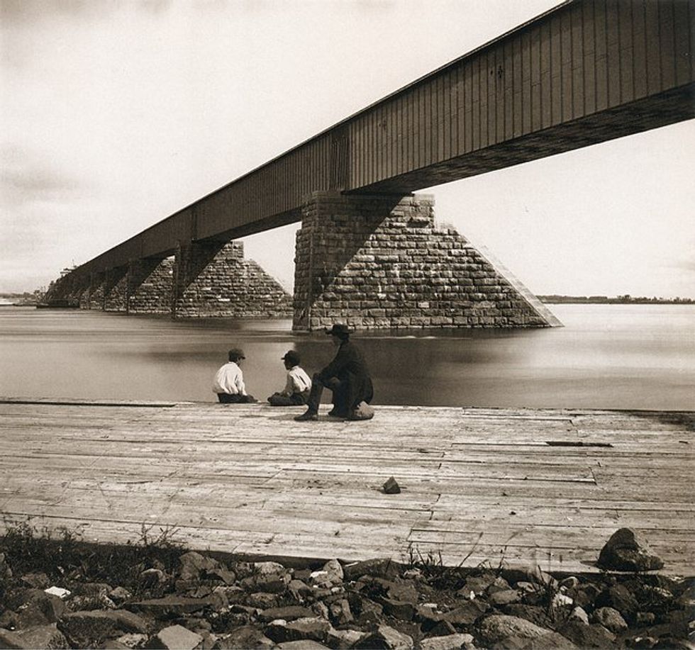 \u200bThe newly-constructed Victoria Bridge in 1859, with three people sitting by the water. The bridge is made of steel box girders.