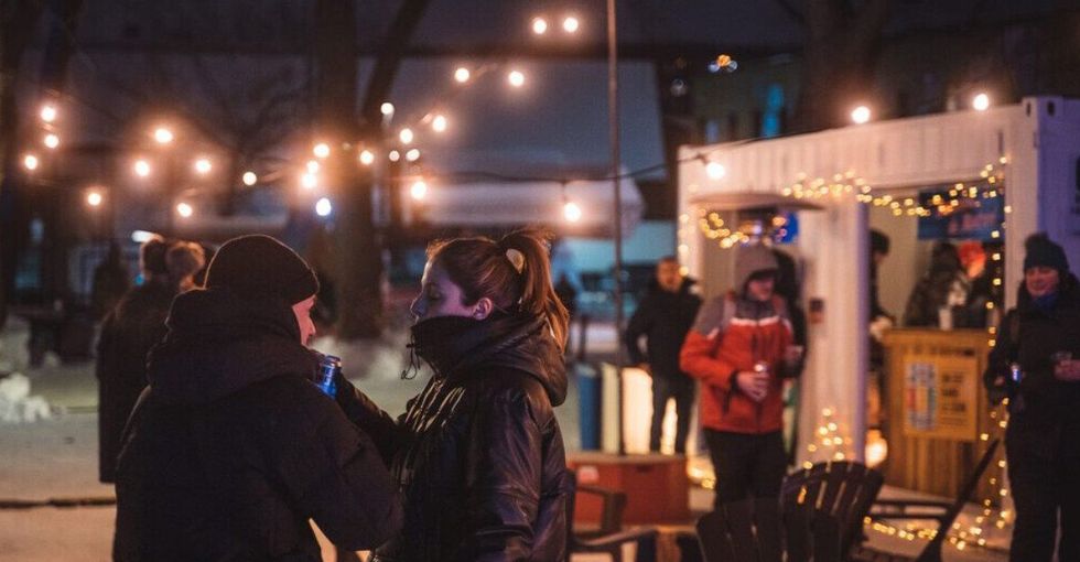 \u200bTwo people in winter coats chat under fairy lights, while others line up for drinks at an outdoor bar.