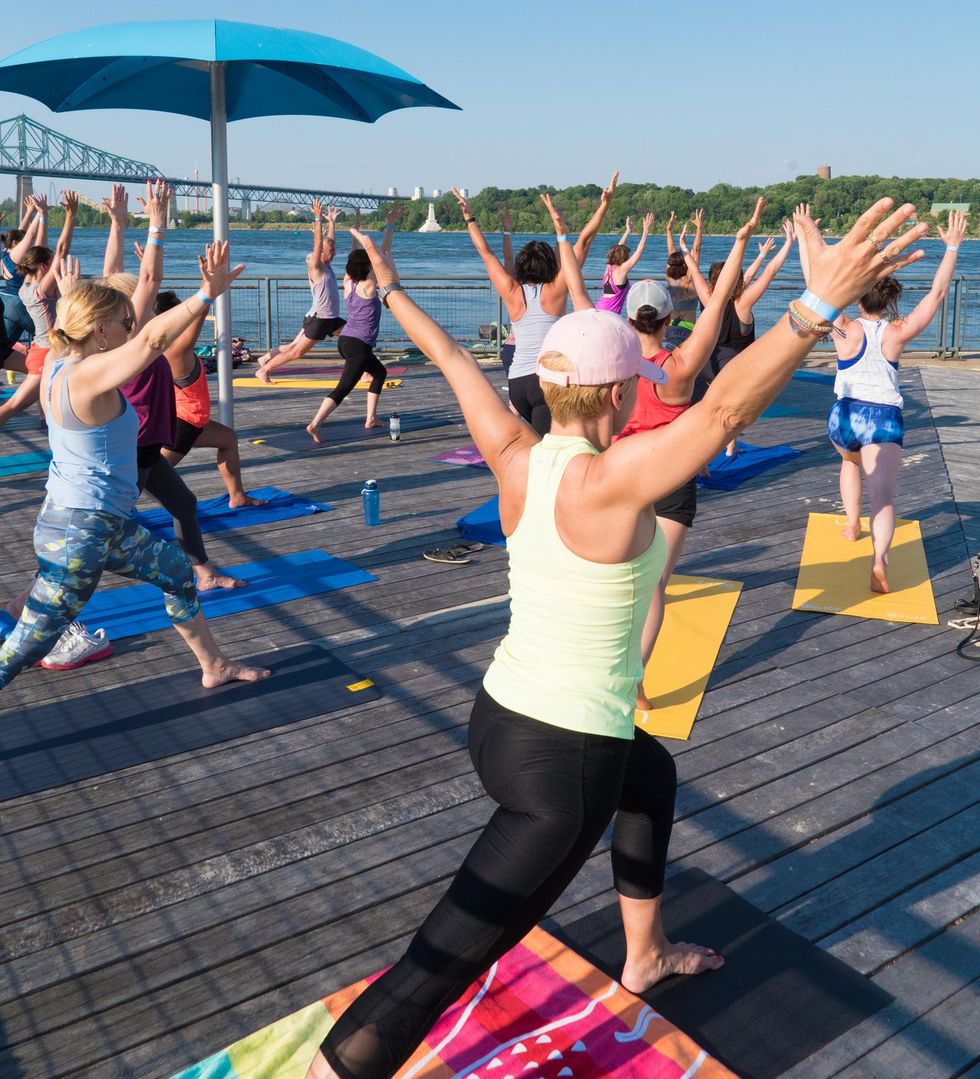 \u200bYoga class participants with one leg forward and arms raised face toward \u00eele Sainte-H\u00e9l\u00e8ne from the Clock Tower Beach boardwalk.