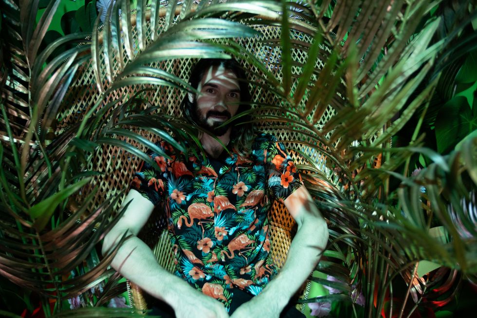 Will wearing a flamingo and hibiscus collared shirt sits in a wicker throne behind gold-tinted tropical plants.